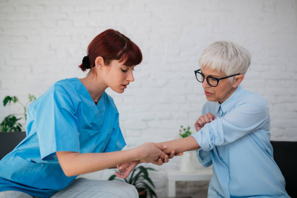 Young adult female nurse in a home visit of an elderly female patient, examining her hand and listening to her patient complaints while she is describing the symptoms to her